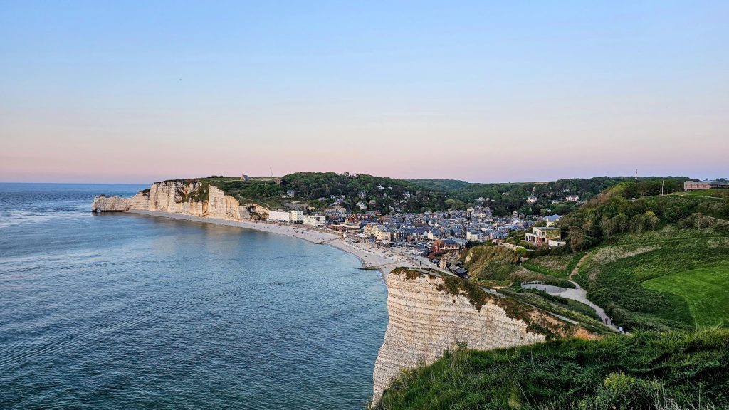 Vista panorâmica de Étretat, na Normandia, com a praia de seixos, a vila costeira e as falésias de giz branco ao entardecer.